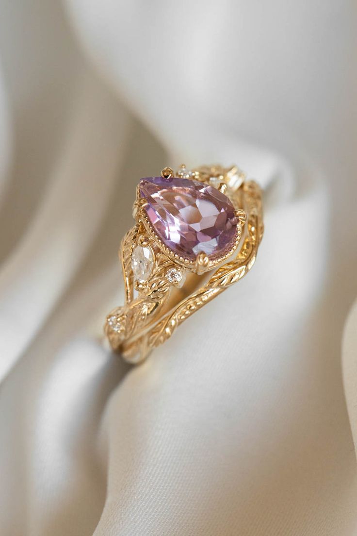 a fancy ring with a pink stone in the center on a white satin material background