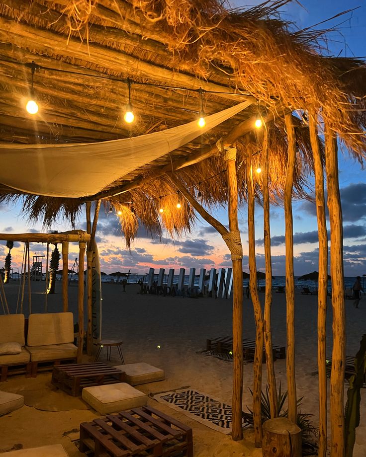 an outdoor seating area on the beach with lights hanging from it's roof and palm trees in the background