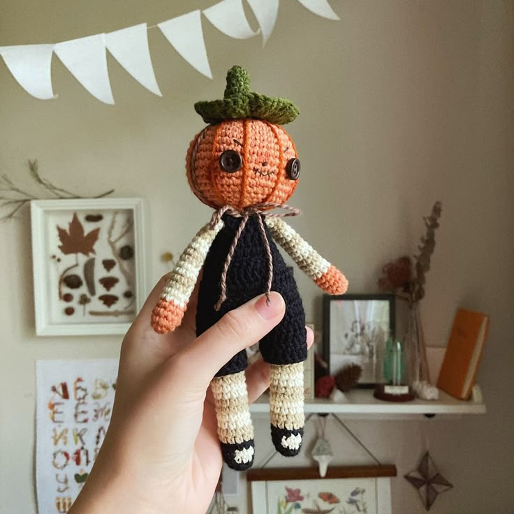 a hand holding a small crocheted pumpkin doll in front of a wall with bunting