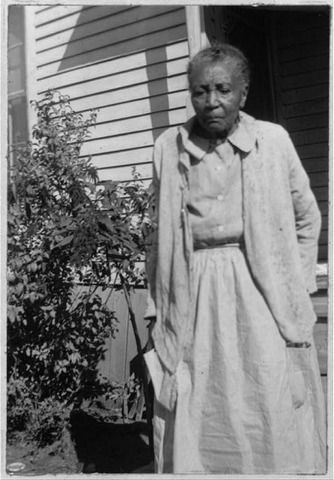 an old black and white photo of a woman standing in front of a house next to a tree