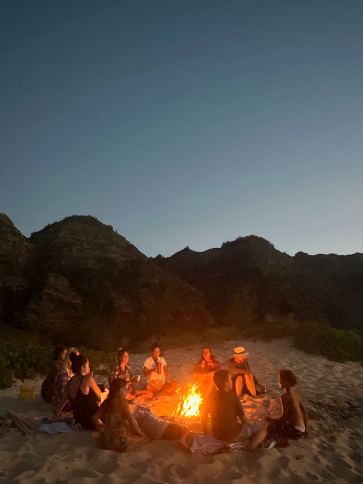 a group of people sitting around a campfire on the beach at night with mountains in the background