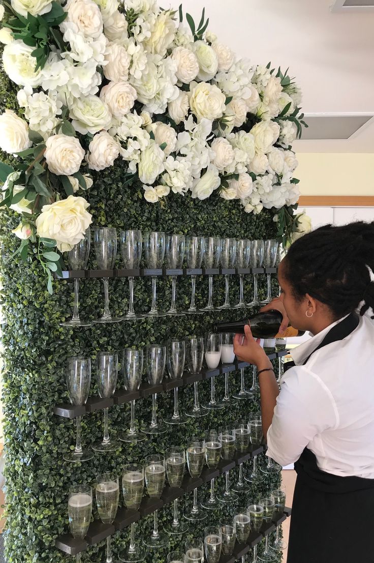 a woman standing in front of a wall filled with wine glasses and white flower arrangements