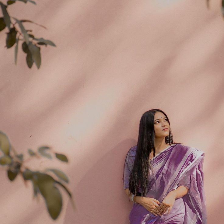 a woman in a purple sari leaning against a pink wall with her hands on her hips