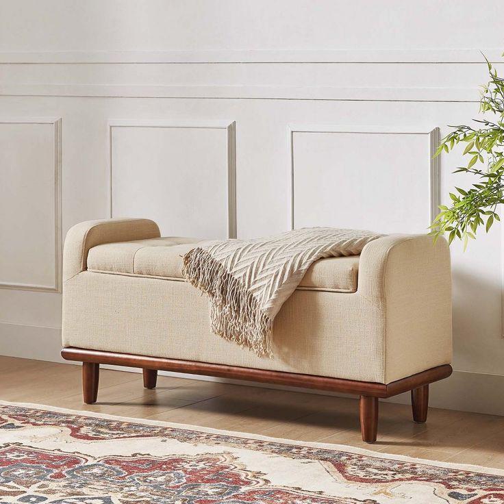 a beige bench with a blanket on it in front of a white wall and rug