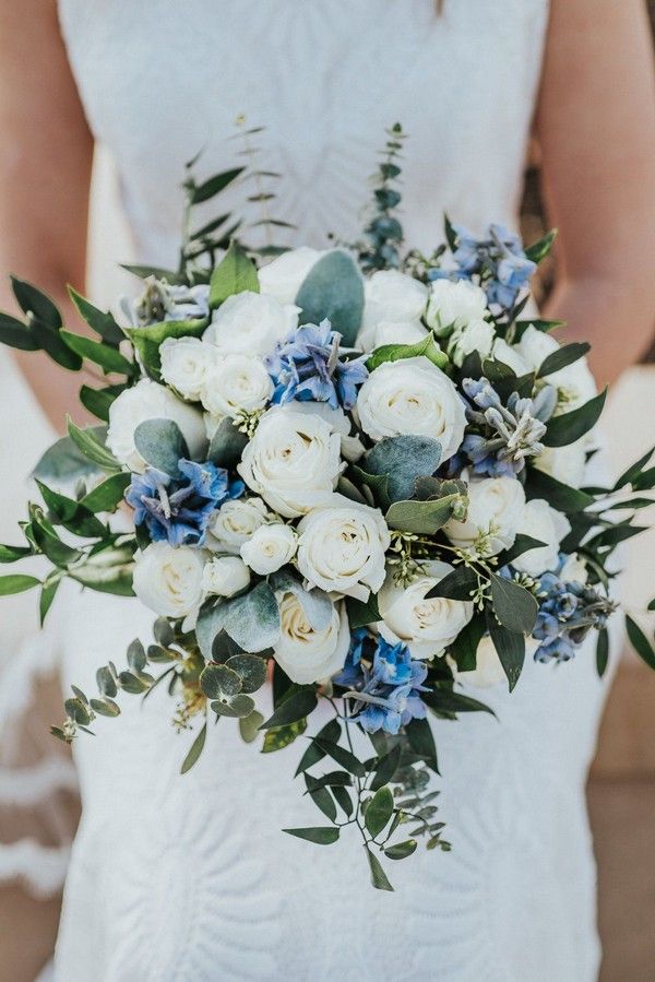 a bridal holding a bouquet of white roses and blue flowers with greenery on the side