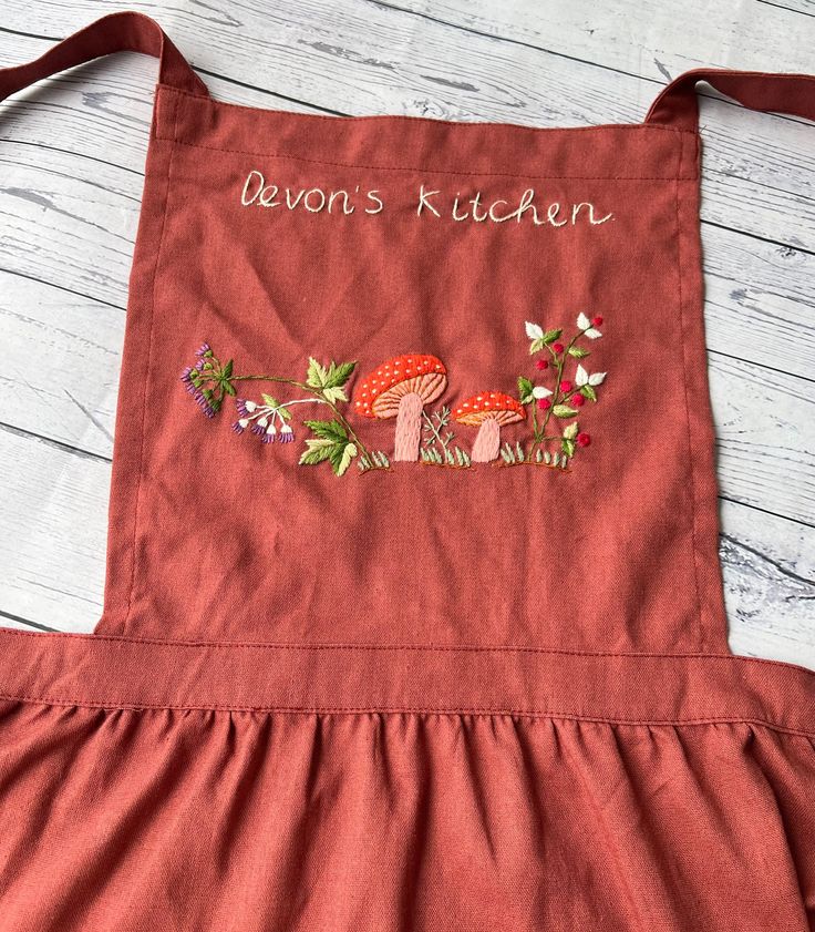 a red apron with embroidered mushrooms and flowers on the front that says baron's kitchen