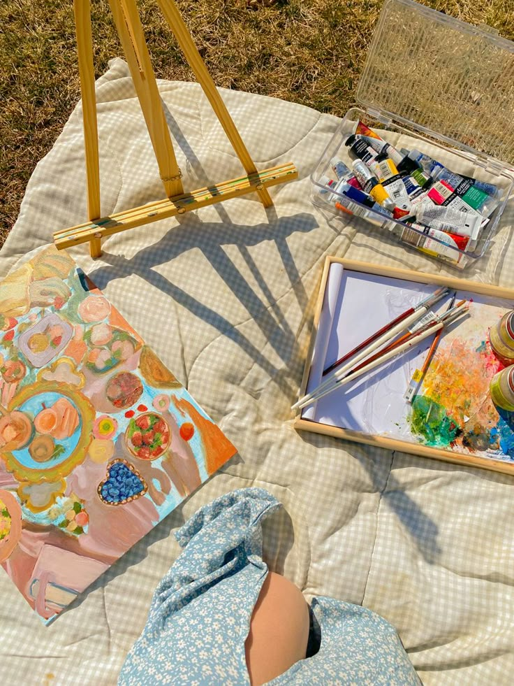 an artist's easel with paint and watercolors sitting on a blanket