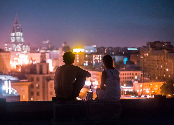 two people are sitting on a ledge looking at the city lights and skyscrapers in the distance