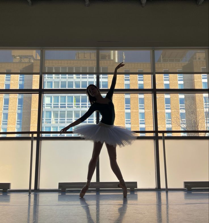 a young ballerina is posing in front of a large window with her arms outstretched