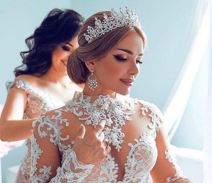 a woman wearing a wedding dress and tiara in front of another woman with her hands on her chest