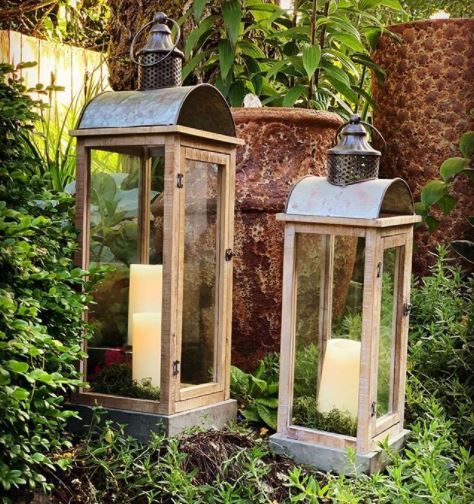 two lanterns are sitting in the grass near some bushes and plants, one is lit with candles