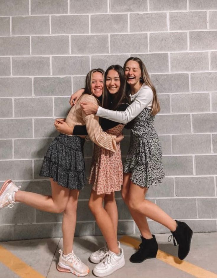 three girls hugging each other while standing in front of a brick wall with their arms around one another