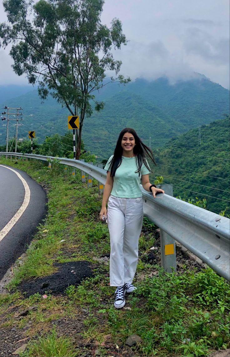 a woman standing on the side of a road next to a lush green hillside with mountains in the background