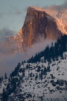 a mountain covered in snow and clouds with trees on the side at sunset or dawn