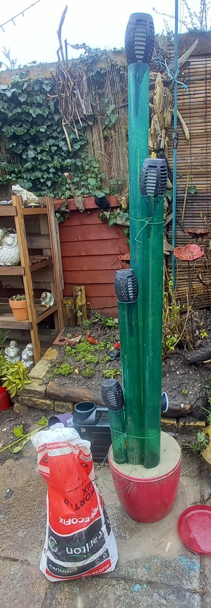 an outdoor garden area with various plants and containers on the ground, including a large green water fountain