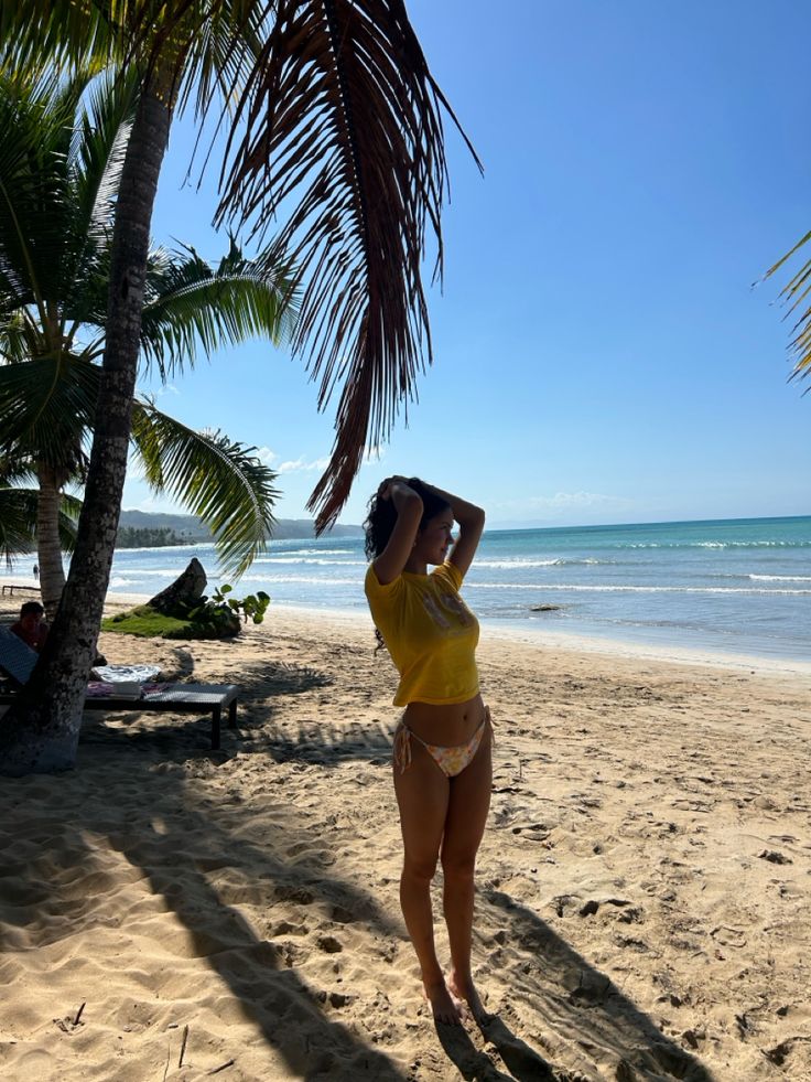 a woman standing on top of a sandy beach under a palm tree next to the ocean