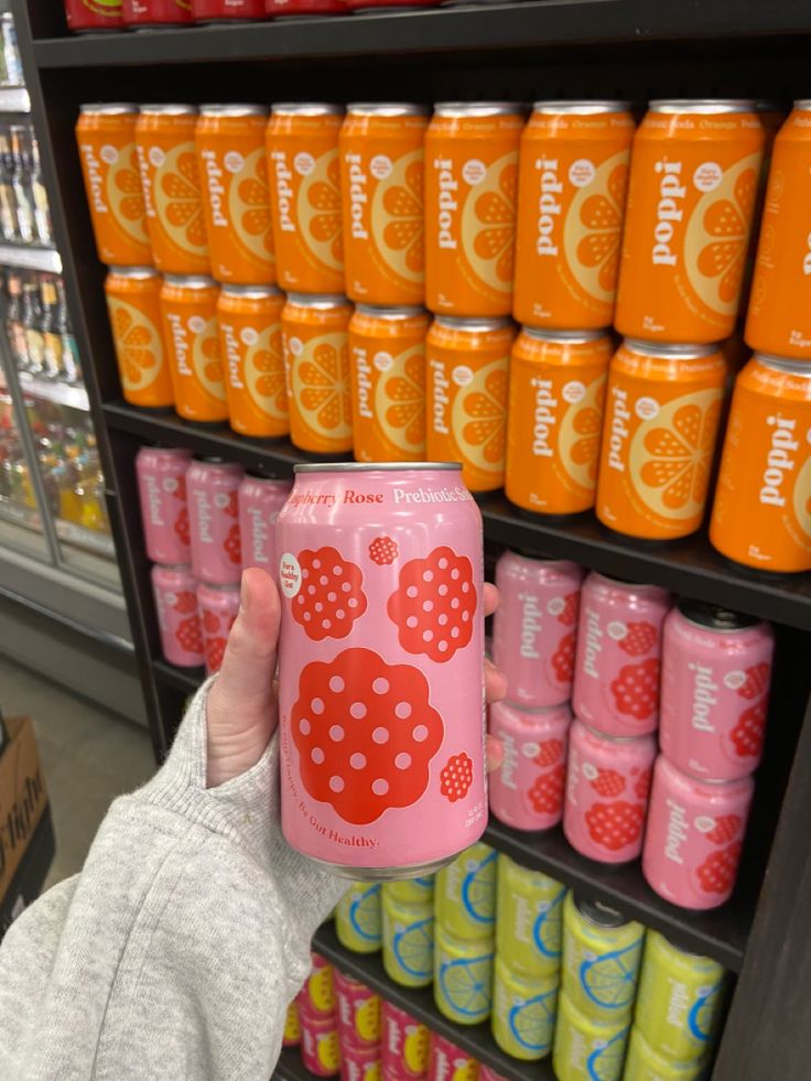 a person holding up a pink and orange can in front of a shelf of soda cans