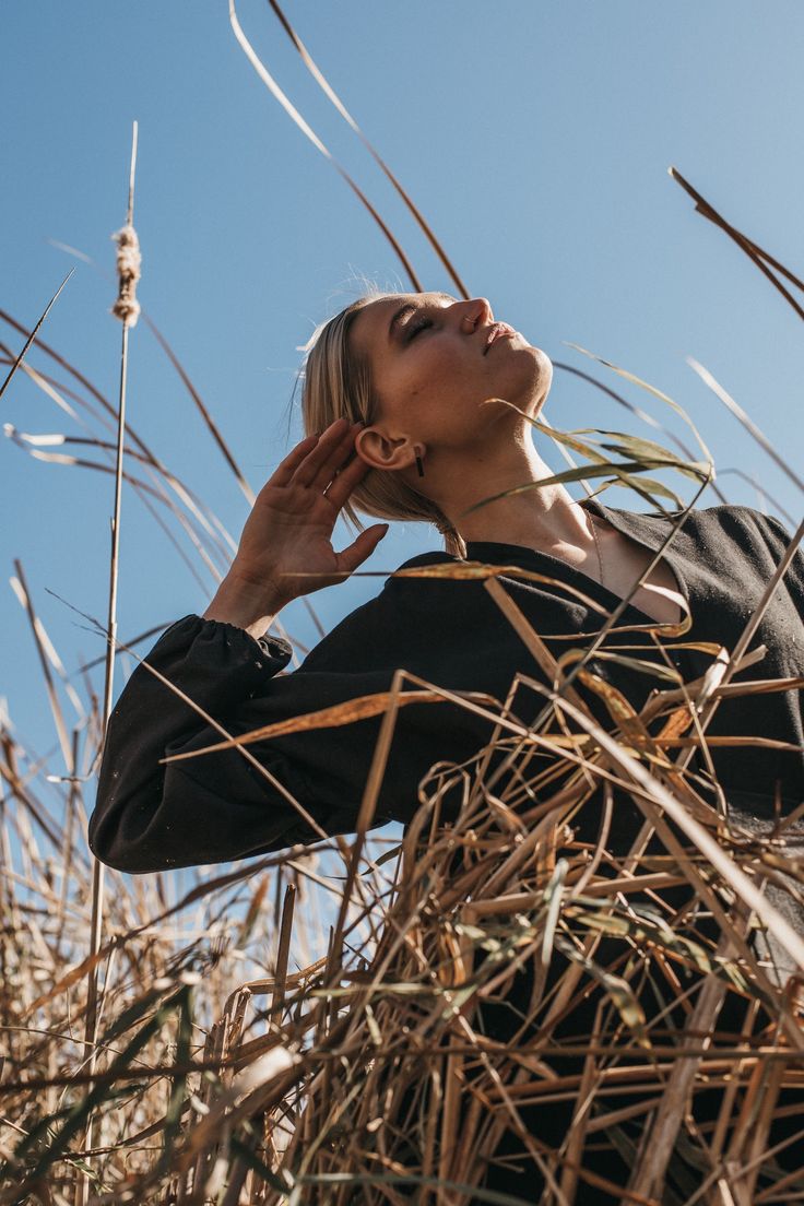 a woman standing in tall grass with her hands to her ear and looking up into the sky