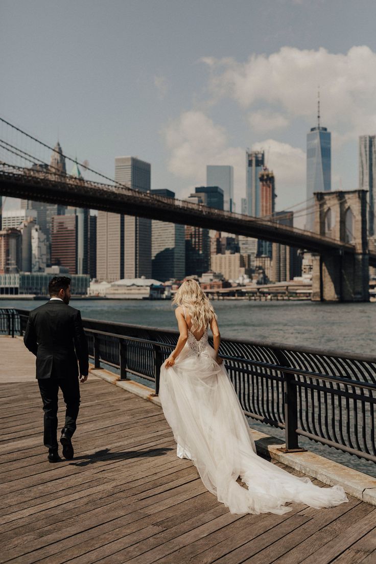 a bride and groom walking on a pier in front of the brooklyn bridge