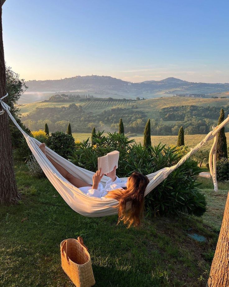 a woman laying in a hammock reading a book on top of a lush green hillside