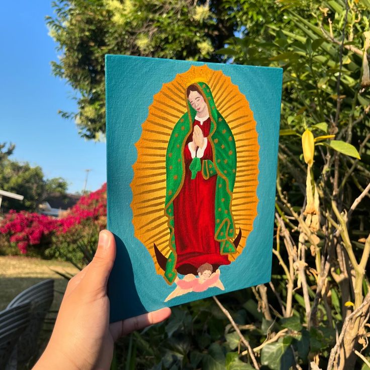 a hand holding up a painting of the virgin mary in front of some trees and bushes