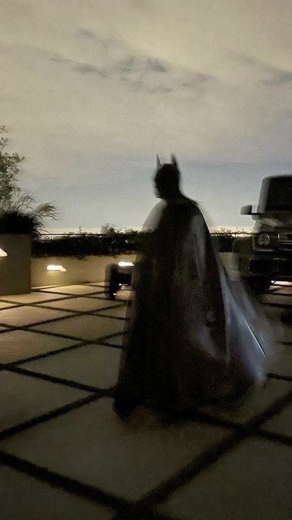 a man in a cape is walking down the street at night with his car behind him