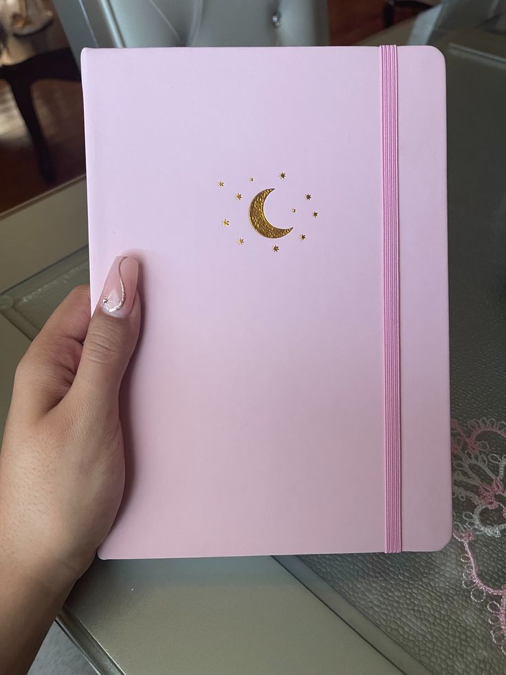 a hand holding a pink notebook with a gold moon on it