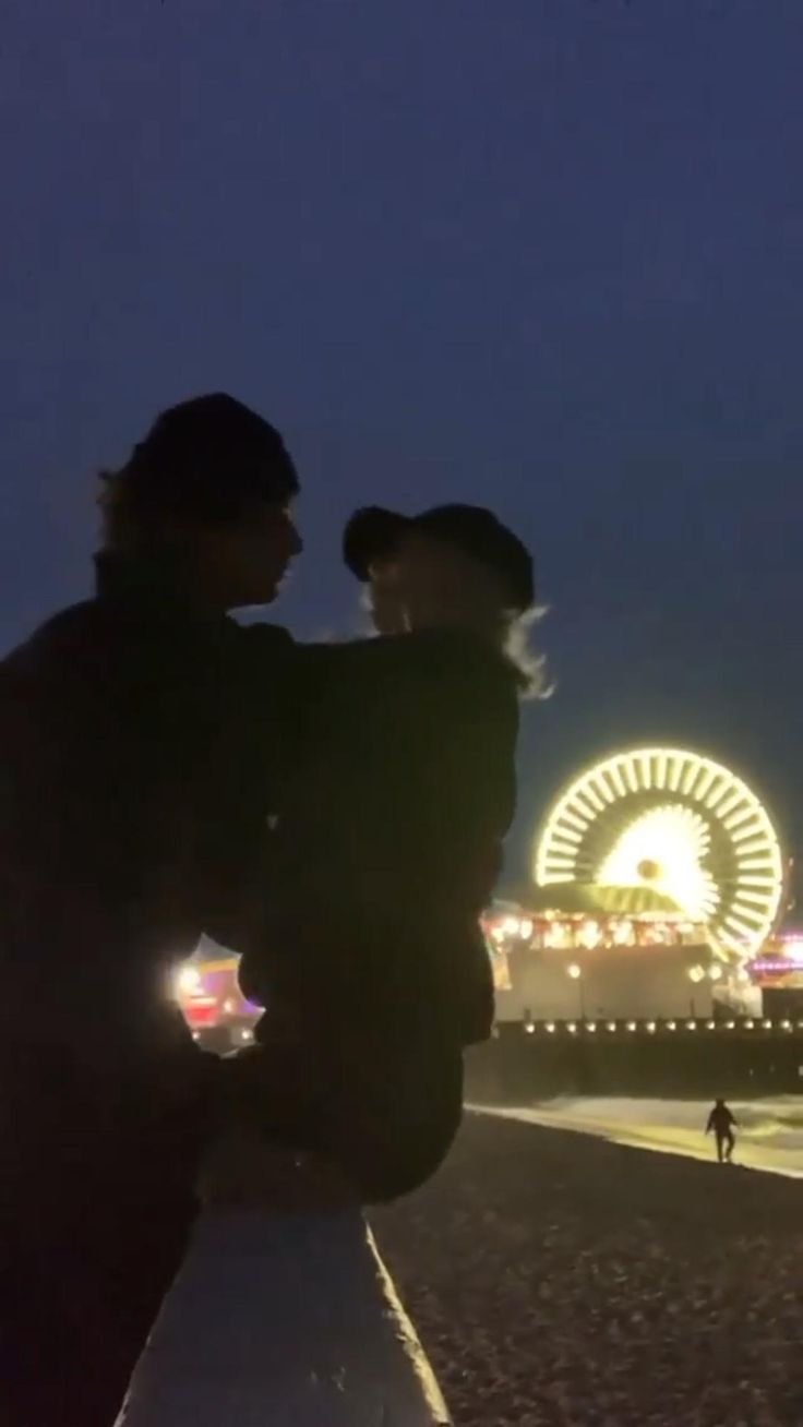 two people standing next to each other in front of a ferris wheel at night time