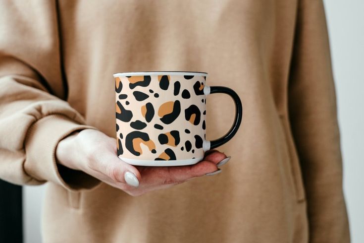 a woman holding a leopard print coffee mug in her hands and wearing a tan sweater