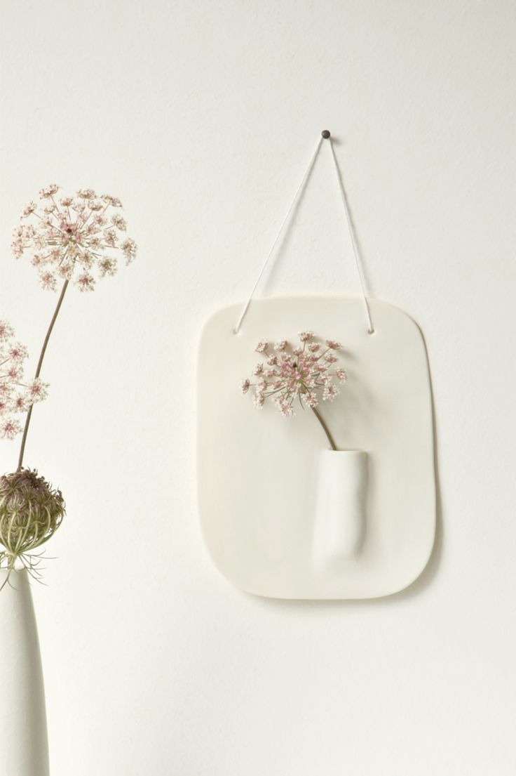 two white vases with flowers in them next to a wall hanging on the wall