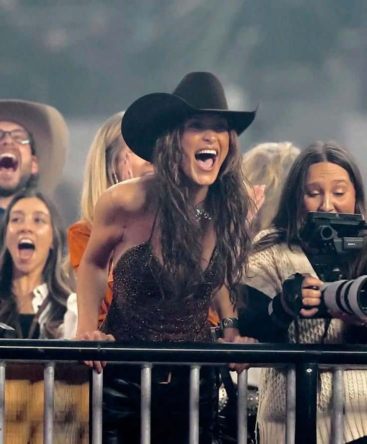 a woman in a cowboy hat is holding a camera and singing into the crowd with her mouth wide open