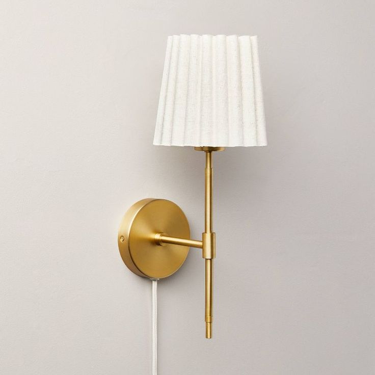 a gold wall light with a white shade on the side and a cord attached to it