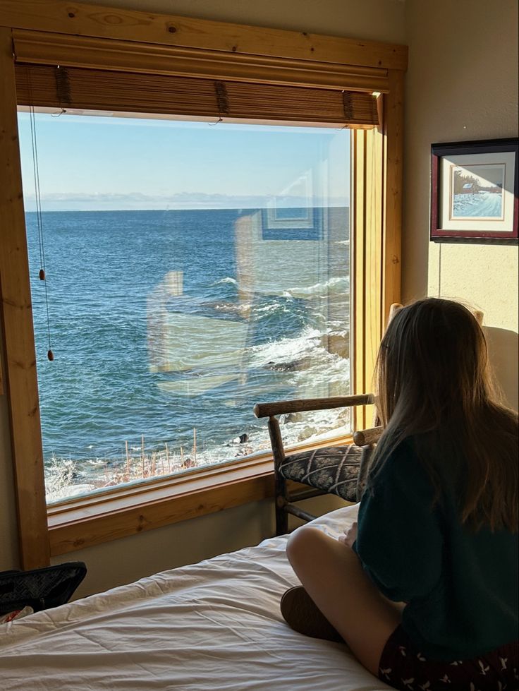 a woman sitting on top of a bed in front of a window looking out at the ocean