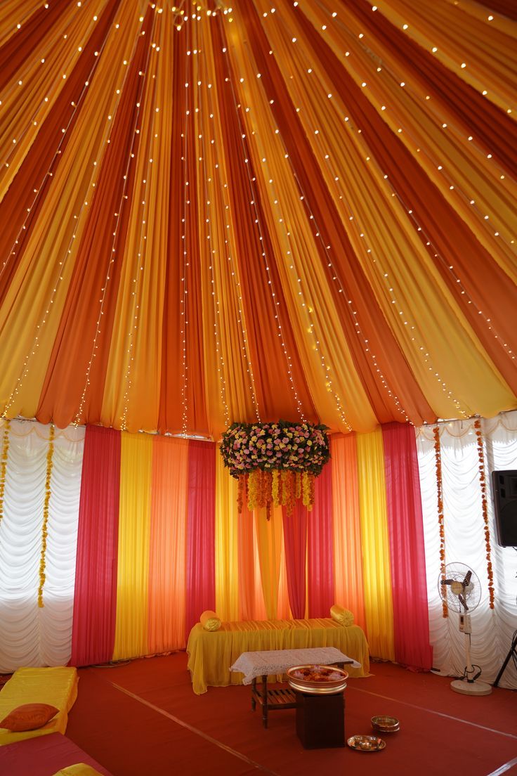 an orange and yellow tent with lights on the ceiling, decorated for a wedding ceremony