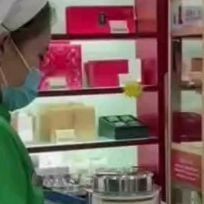 a woman wearing a surgical mask in front of a refrigerator filled with cheese and other items