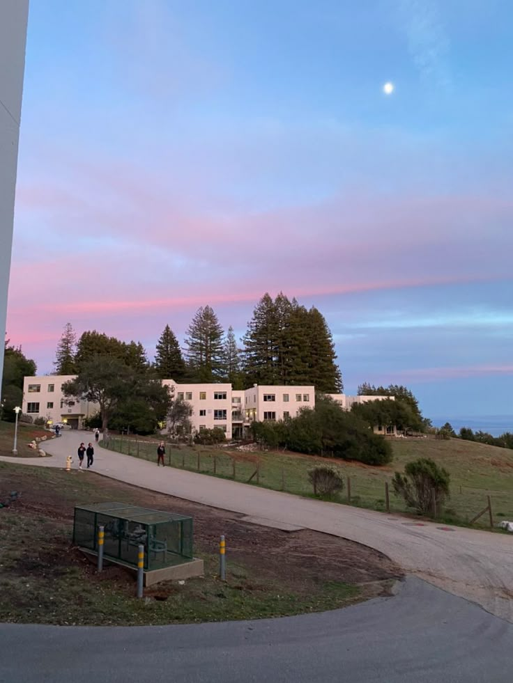 people are walking on the path near some buildings and trees at sunset or sunrise time