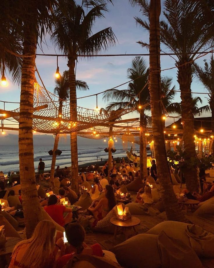 many people are sitting on the beach under palm trees at night with lights strung over them