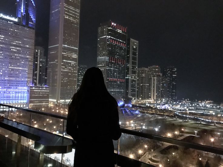 a woman standing in front of a window overlooking a city skyline at night with buildings lit up