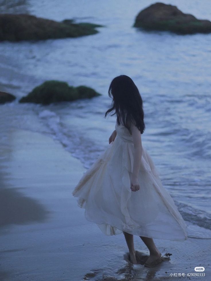 a woman in a white dress walking on the beach at dusk with her hair blowing in the wind