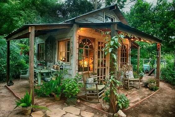 a small wooden cabin in the middle of some trees and bushes with lights on it