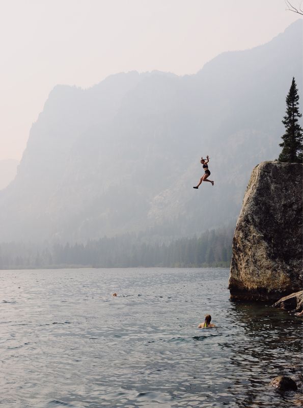 a person jumping into the water from a cliff in front of some mountains and trees