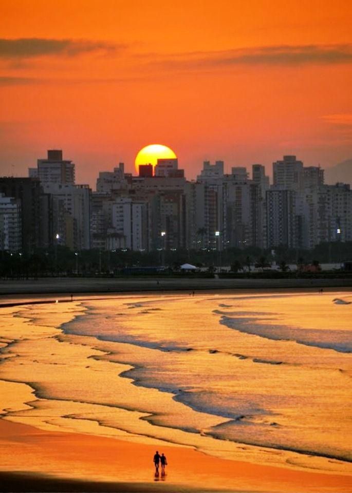 two people walking on the beach in front of an orange sky with buildings behind them