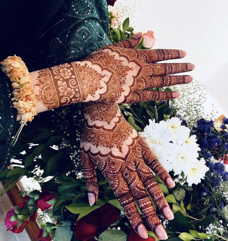 two hands with henna tattoos on their palms and flowers in the foreground, against a white background