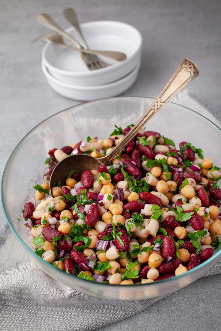 a glass bowl filled with beans and greens next to a white bowl full of silverware