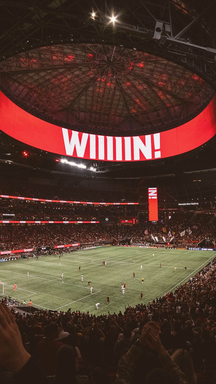 a stadium filled with people watching a soccer game on a big screen in the middle of the room