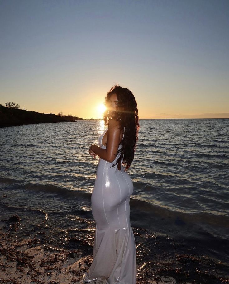 a woman in a white dress standing on the beach at sunset with her back to the camera