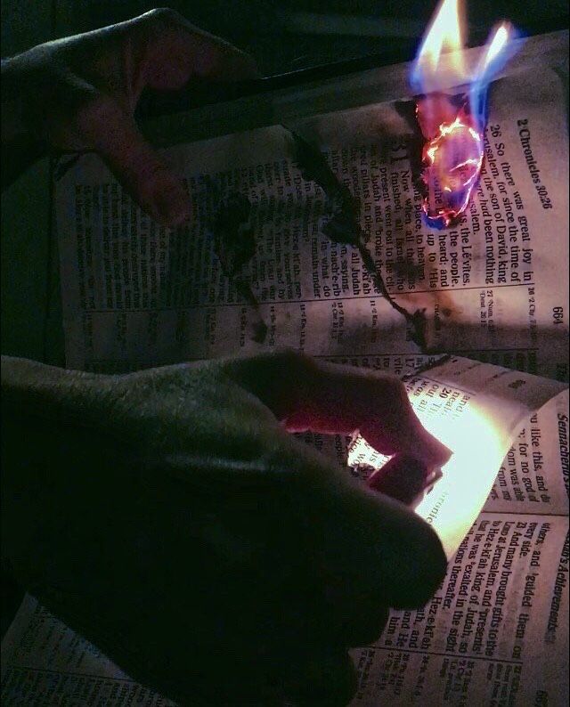 two hands holding a lit candle on top of an open newspaper with flames coming out of it