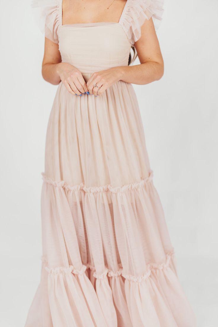 This dress will let your inner princess shine through! Meet the Bella, the dreamy, tulle-layered ballgown we're all obsessing over. This beauty is light as air and destined for the dance floor; from its square neckline and ruffled cap sleeves, to its voluminous multi-tiered tulle skirt, the Bella will capture everyone's attention when you walk in the room. Just don't forget your glass slippers! Available in four colorways. See fabric swatch for true "Peachy Nude" color. FIT: Runs true to size. Y Pink Tiered Dress With Voluminous Skirt, Pink Tiered Voluminous Dress, Pink Voluminous Tiered Dress, Flowy Ruffled Tulle Dress, Feminine Tiered Maxi Dress For Party, Feminine Tiered Skirt Maxi Dress For Party, Flowy Tulle Dress With Ruffled Skirt, Tiered Tulle Skirt Dress, Blush Tulle Dress With Ruffles