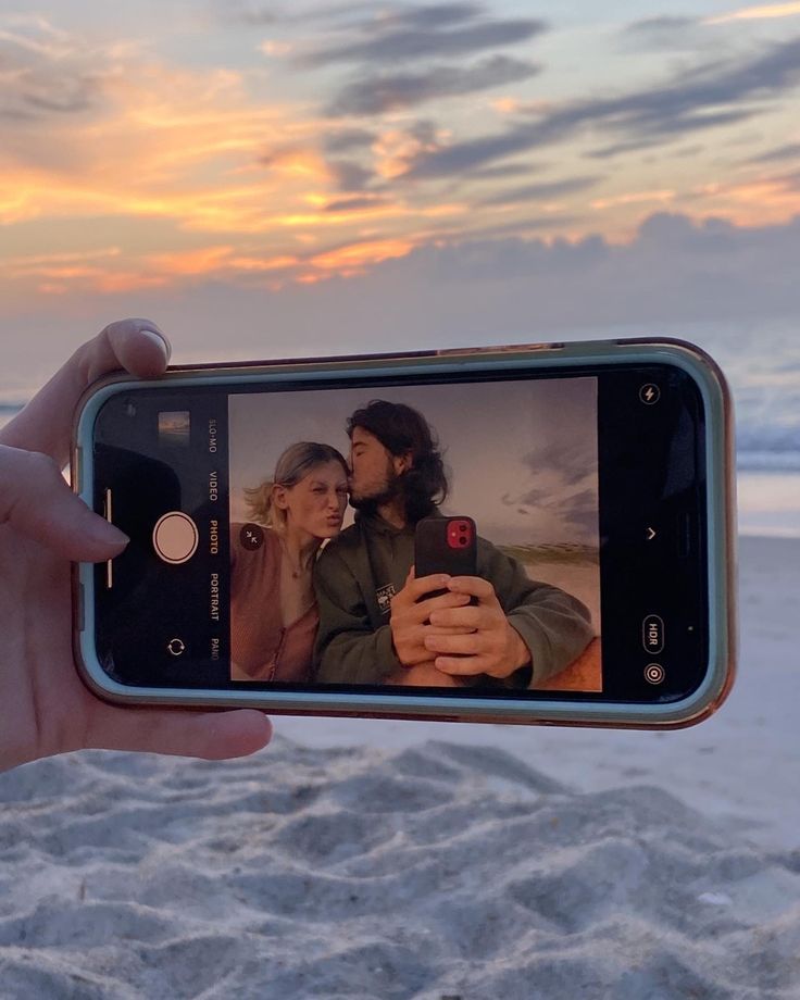 a person holding up a cell phone to take a photo on the beach at sunset