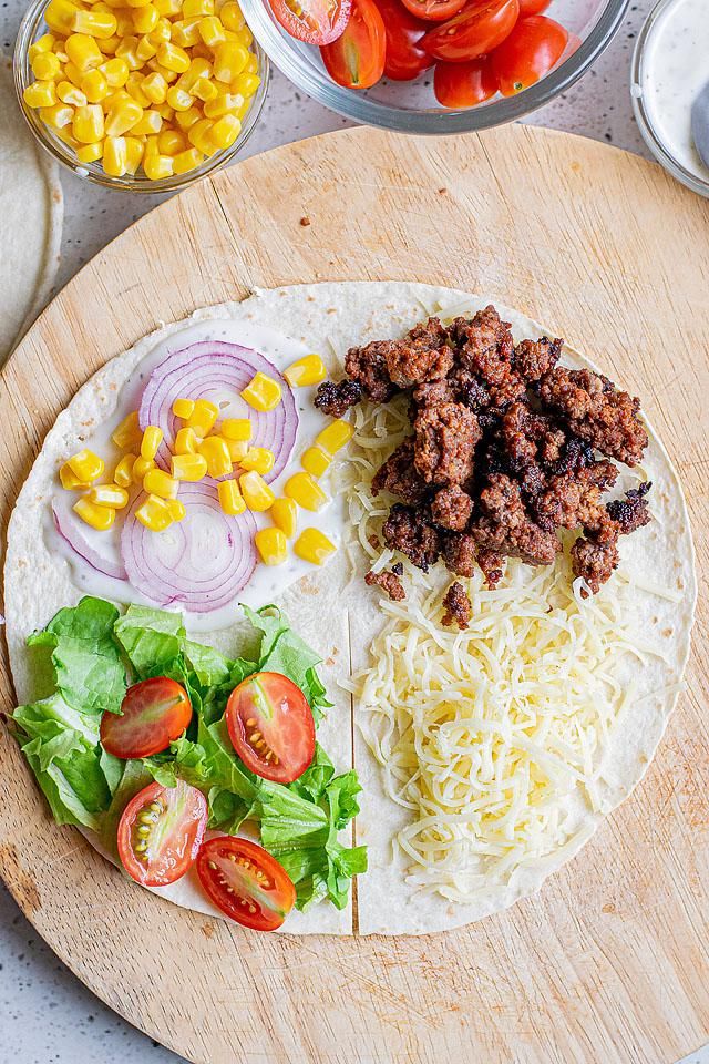 a tortilla topped with meat, cheese and vegetables on a wooden cutting board